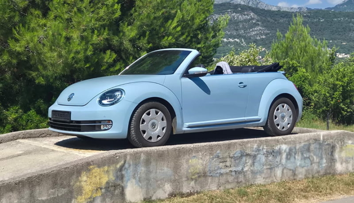 Ein hellblaues Volkswagen-Käfer-Cabrio steht auf einer Betonplattform, im Hintergrund sind Bäume und Berge zu sehen.